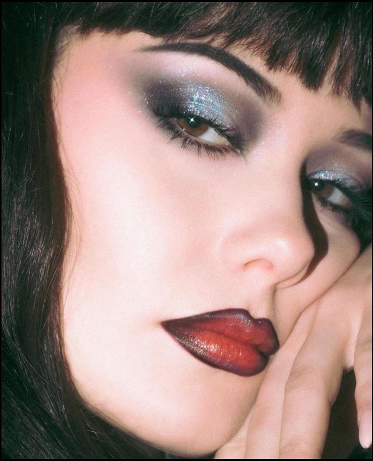 Close-up of a woman's face with dramatic makeup. Her dark hair is partially visible, and her eyes are accentuated with shimmering silver eyeshadow, dark eyeliner, and dark red lipstick. Her eyebrows are neatly trimmed, and her hand rests gently on her chin.