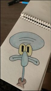A drawing of Squidward from SpongeBob SquarePants is shown on a spiral-bound sketchbook. The drawing features Squidward's face with large, round eyes, a prominent nose, and a long blue tube extending downwards. A brown, pouch-like shape is positioned at the bottom of the drawing. Two colored pencils or markers are visible in the background.