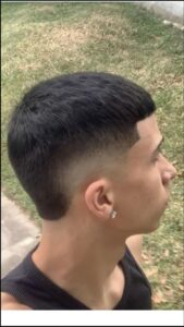 A young man with a short, dark haircut and a side-swept fringe wears a dark sleeveless top and a small stud earring. He is positioned against a blurred background of grass and a paved area.