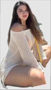 A young woman with long brown hair wears a white, off-the-shoulder, knitted dress. She is seated against a light background and looks directly at the camera with a slight smile.