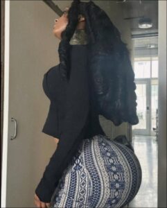 A woman with long black hair, wearing a black top and patterned skirt, stands in a hallway, looking upwards. She is positioned in front of a mirror reflecting a doorway and a glimpse of another room.