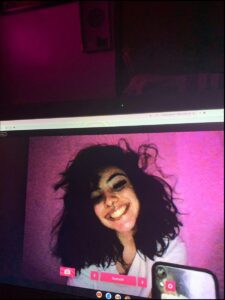 A computer screen displays a selfie of a young woman with dark hair, smiling and holding a smartphone. The background is a pinkish-purple hue.