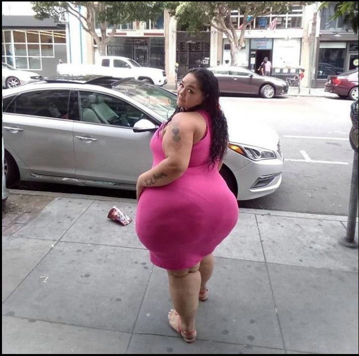 A woman wearing a pink dress stands on a city sidewalk next to a parked silver car. She has tattoos on her left arm and wears sandals. A crumpled piece of paper lies on the ground near her feet. Buildings and parked cars are visible in the background.