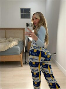 A young woman takes a mirror selfie in a bedroom, wearing patterned pajama pants featuring Corona Extra beer designs. She holds a smartphone and wears a light blue long-sleeved shirt. A bed with a light-colored comforter and a window are visible in the background.