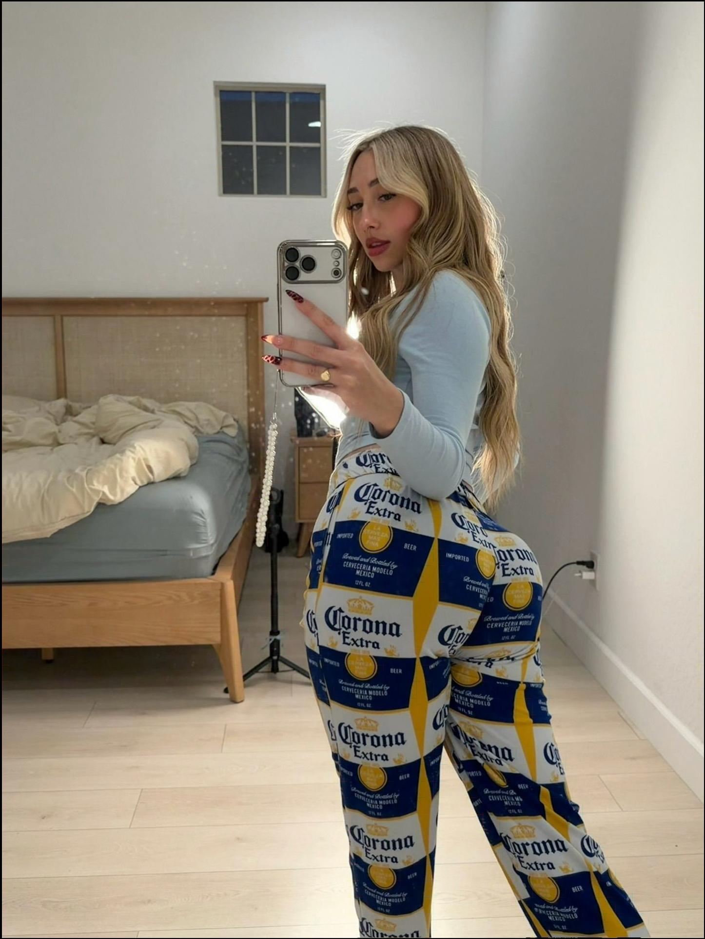A young woman takes a mirror selfie in a bedroom, wearing patterned pajama pants featuring Corona Extra beer designs. She holds a smartphone and wears a light blue long-sleeved shirt. A bed with a light-colored comforter and a window are visible in the background.
