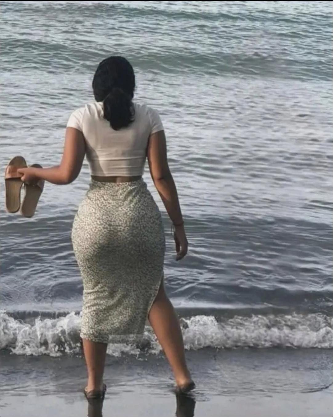 A woman stands in shallow water at the edge of a beach, facing the ocean. She wears a white top and a light green floral skirt, holding sandals in her left hand.