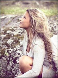 A young woman with long, light blonde hair sits crouched on a rocky surface, looking upwards. She wears a light gray cardigan and a white garment underneath. The background is out of focus, showing a grassy area.