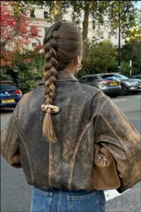 A woman with a braided hairstyle wears a distressed brown leather jacket and blue jeans, carrying a brown purse. She is standing on a city street with parked cars and trees in the background.