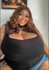 A woman with voluminous brown hair wears a black top and denim shorts. She smiles at the camera against a textured white wall with a framed picture.