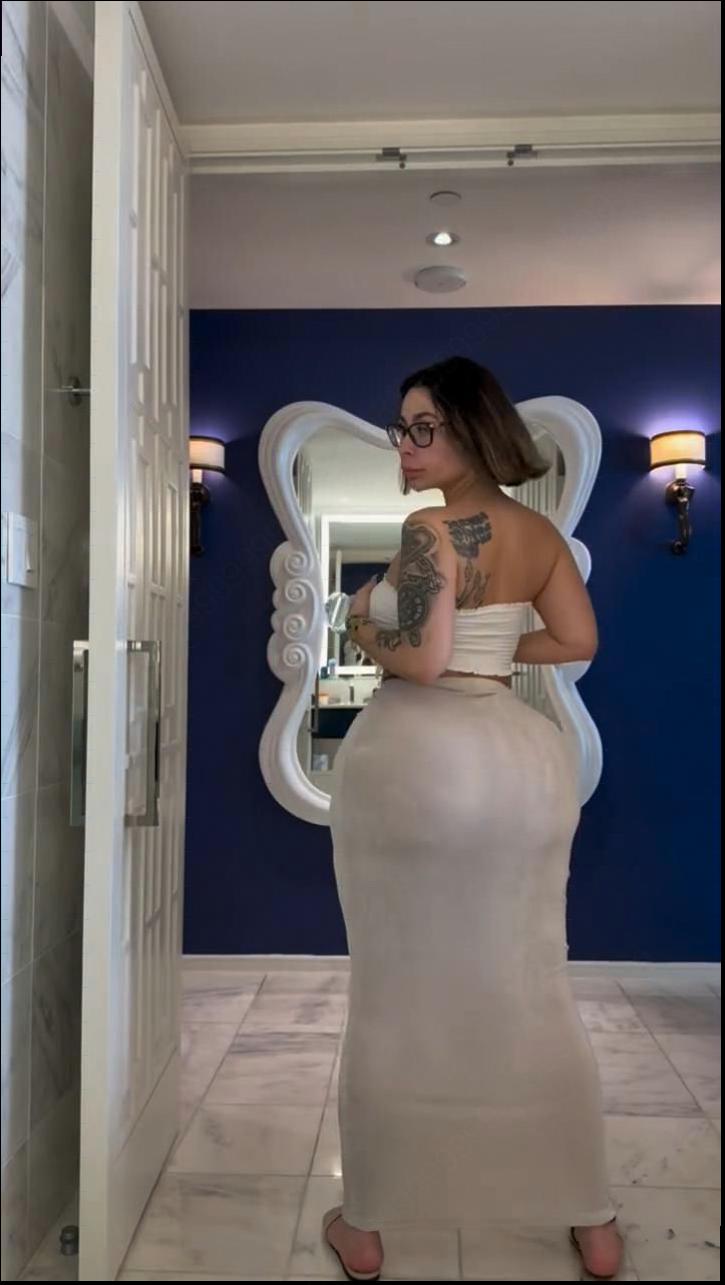 A woman with tattoos on her back and arms stands in front of a mirror in a bathroom. She wears a white top and a long skirt, and her reflection is visible in the mirror. The bathroom features dark blue walls, white trim, and a marble floor.