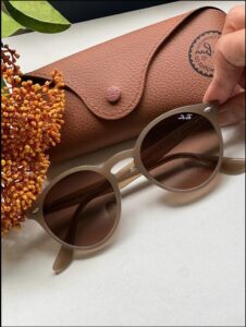 A pair of round sunglasses with brown lenses rests on a white surface next to a brown leather Ray-Ban case. A dried floral arrangement is partially visible in the lower left corner.