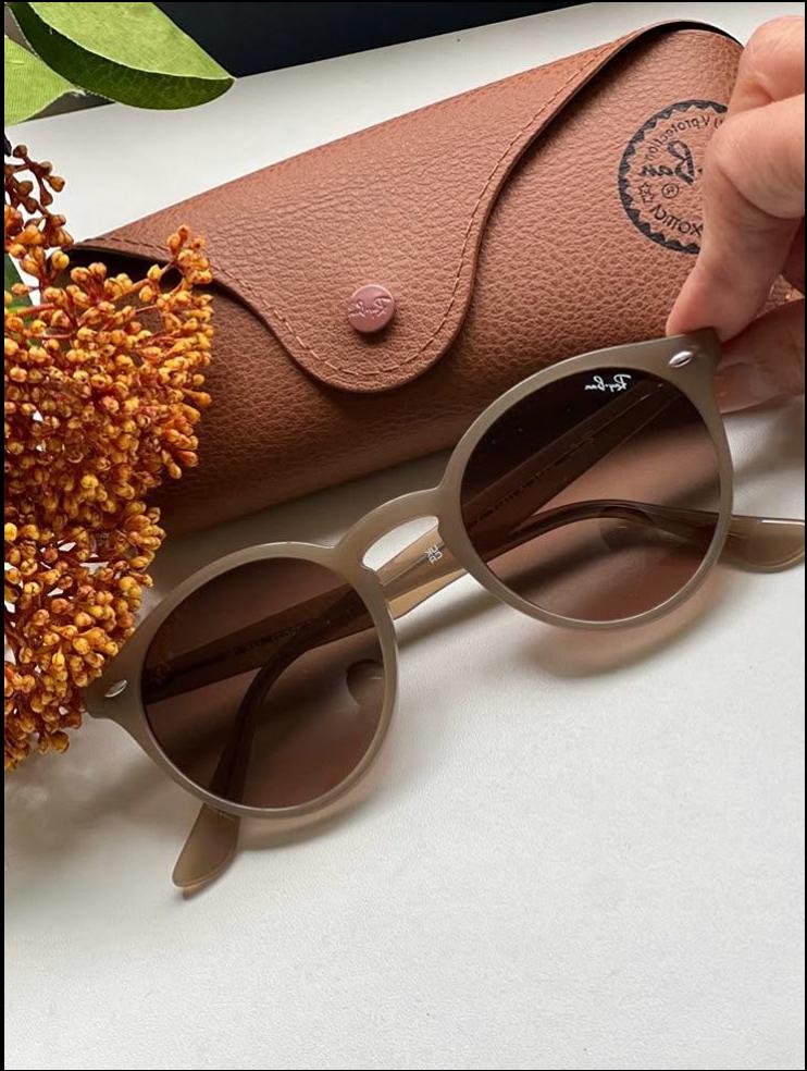 A pair of round sunglasses with brown lenses rests on a white surface next to a brown leather Ray-Ban case. A dried floral arrangement is partially visible in the lower left corner.