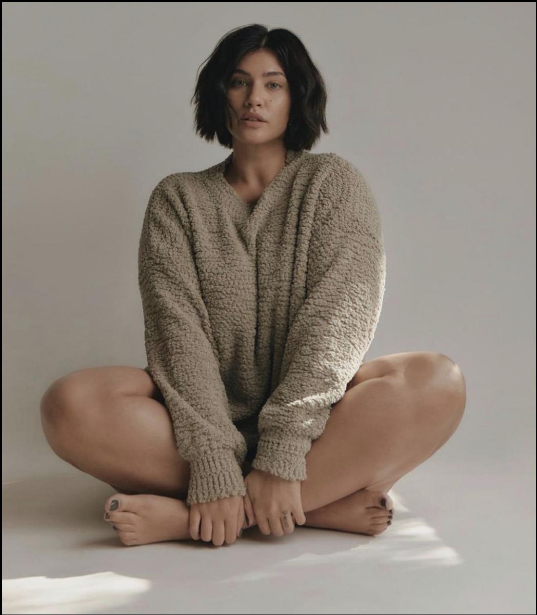 A woman sits cross-legged on a white surface, wearing a light brown knit sweater. Her bare feet are visible beneath her knees, and she looks directly at the camera.
