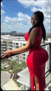 A woman in a red dress stands on a balcony overlooking a cityscape with buildings, trees, and a pool. She is looking over her shoulder towards the camera.