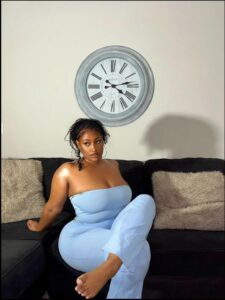 A woman in a light blue strapless jumpsuit sits barefoot on a black couch. She is positioned in front of a wall-mounted clock with a light-colored face and dark Roman numerals. Tan and beige throw pillows are visible on the couch.