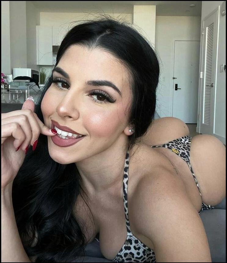 A woman with long dark hair, wearing a leopard-print bikini top and bottoms, lies on her stomach in a room. She is touching her lip with her finger and smiling at the camera.