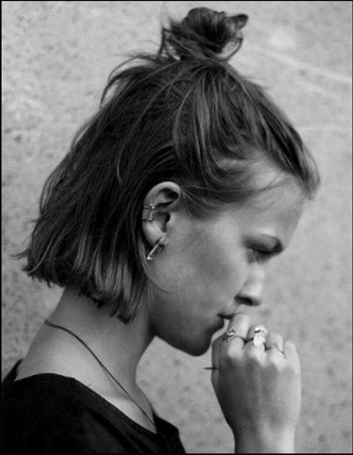 Black and white profile view of a person with short, textured hair styled in a topknot, wearing multiple piercings, including earrings and a necklace. They are touching their face with their right hand and wearing rings on their fingers.