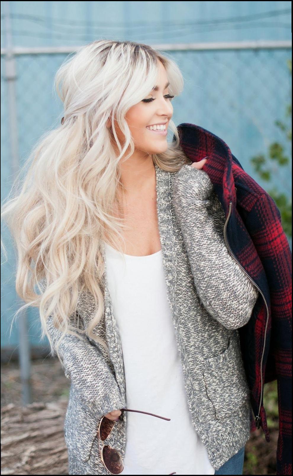 A young woman with long, light blonde hair wears a gray knitted cardigan over a white t-shirt and holds a pair of sunglasses. She stands against a blue-gray chain-link fence with a hint of greenery visible in the background.