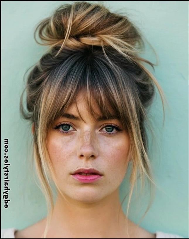 A young woman with light brown hair styled in a loose bun with bangs and loose strands framing her face. She has freckles and light pink lipstick, and her eyes are light blue. The background is a pale teal.