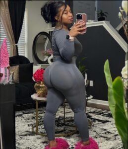 A woman wearing a gray leggings top and pink fuzzy slippers takes a mirror selfie in a living room. She holds a rose-gold phone and wears a watch. A gold coffee table with red flowers, a black couch, and a gold mirror are visible in the background.