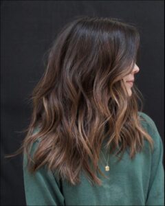 A person with long, wavy hair in shades of brown and gold wears a dark green top and a gold necklace. The hair is styled with loose waves and highlights, falling over their shoulders and partially obscuring their face.