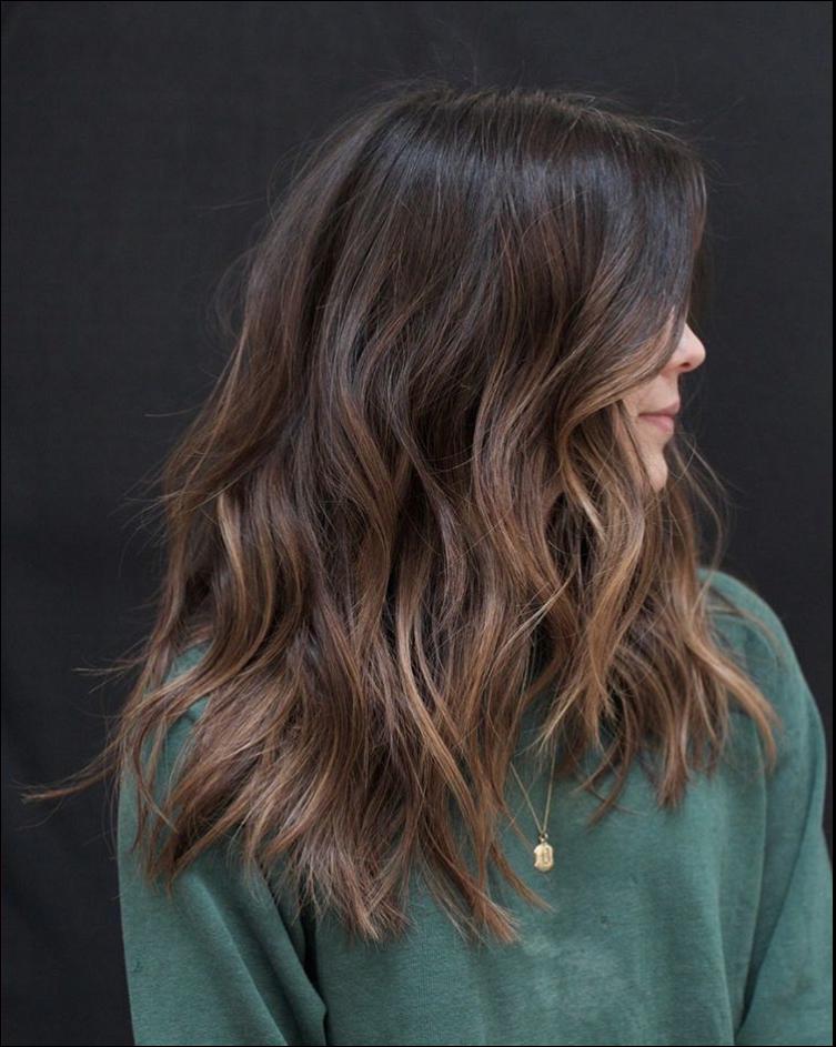 A person with long, wavy hair in shades of brown and gold wears a dark green top and a gold necklace. The hair is styled with loose waves and highlights, falling over their shoulders and partially obscuring their face.