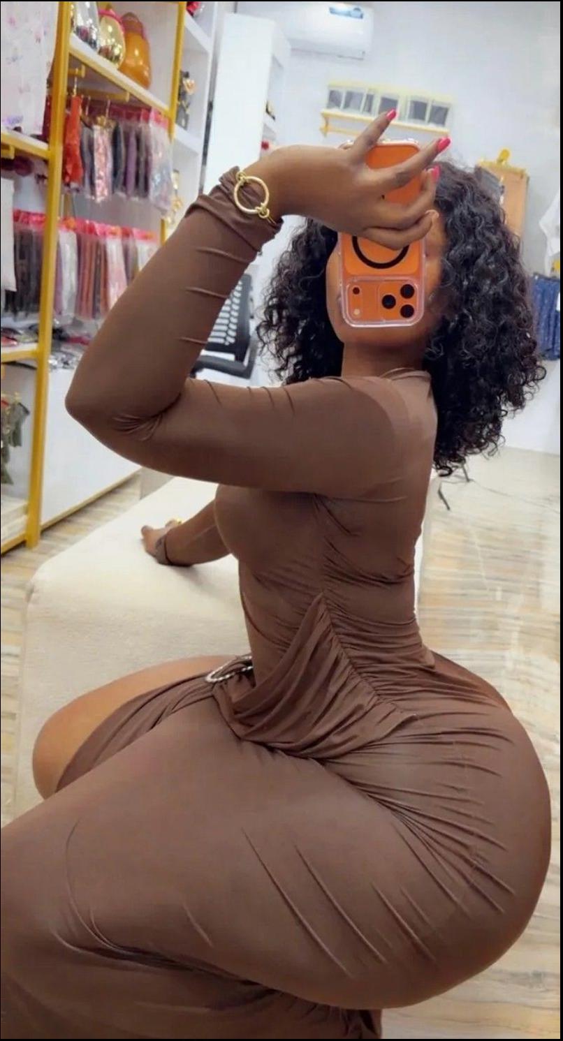 A woman with curly hair, wearing a brown long-sleeved dress and a gold bracelet, takes a mirror selfie in a store. She holds an orange phone in her right hand and looks at the reflection. A display rack with various items is visible in the background.