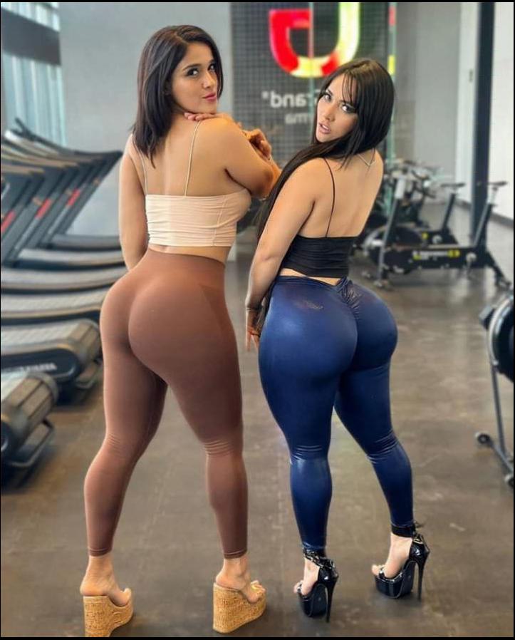 Two women pose in a gym wearing leggings. One wears brown leggings and a light-colored crop top, while the other wears blue leggings and a black crop top. Both wear platform heels. The gym background includes treadmills, stationary bikes, and a large "U" logo.