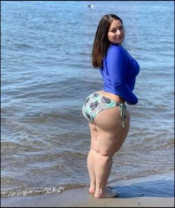 A woman stands on a sandy beach, wearing a blue long-sleeved rash guard and patterned bikini bottoms. She is facing the camera and looking over her shoulder towards the ocean.