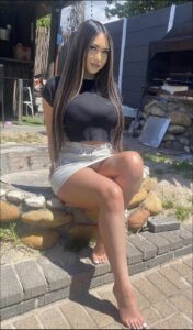 A young woman with long dark hair, wearing a black top and light-colored mini skirt, sits barefoot on a brick and stone patio. She is positioned in front of a partially visible outdoor structure with a grill and fire pit.