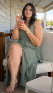 A woman in a green dress sits barefoot on a white bench, holding a drink in a clear glass with a red rim. She wears a bracelet on her left wrist and a necklace. A wooden structure and a partially visible white chair are in the background.