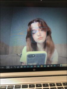 A screenshot shows a young person with long reddish-brown hair using a smartphone in a video call. A blue box overlays their face, indicating a face recognition feature. The laptop screen displays a blurred background.