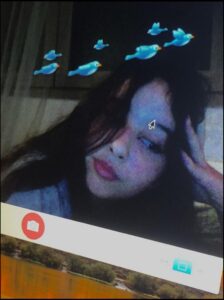 A close-up selfie of a young person with long dark hair, wearing a light-colored top, against a blurred background. Six blue birds are superimposed above the person's head. A camera icon is visible in the lower left corner of the image.