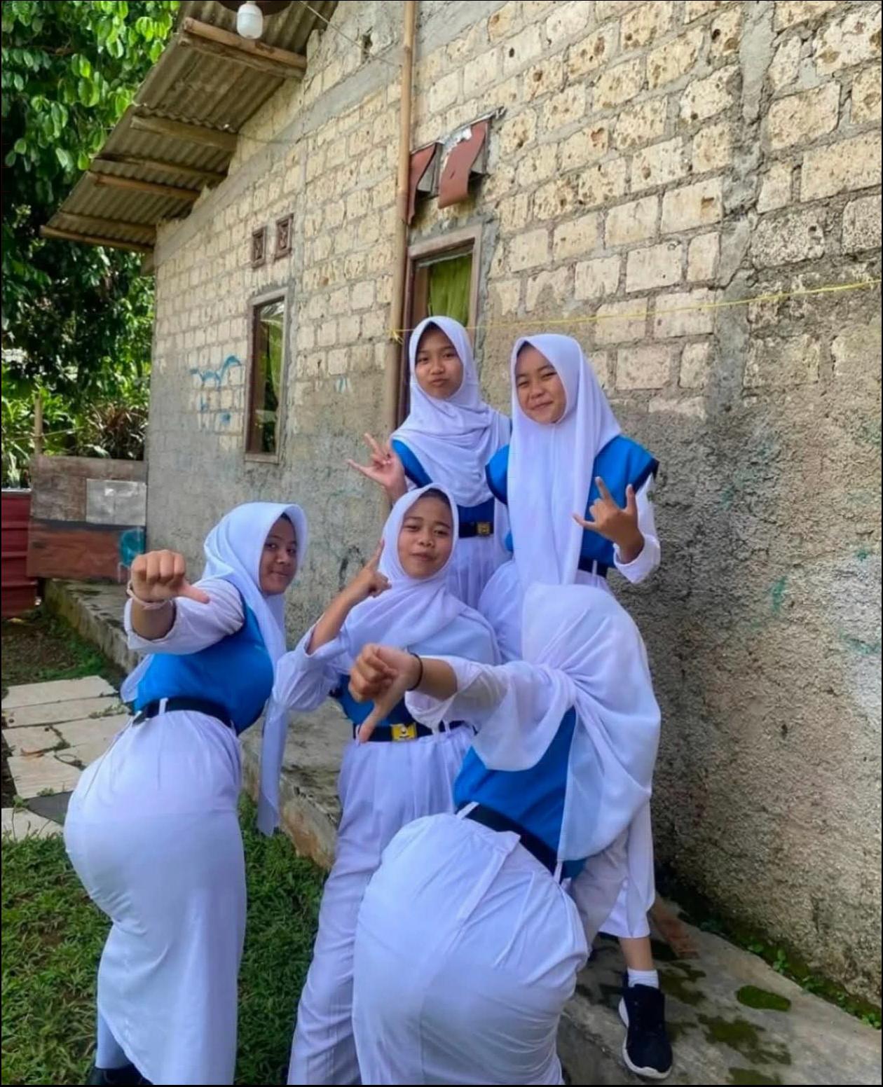 Six girls in school uniforms pose in front of a light-colored building with a corrugated metal roof. They are wearing white skirts and blue tops, with some making hand gestures.