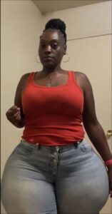 A woman with dark skin and styled hair wears a red tank top and light blue jeans. She is standing against a light-colored wall with a partially visible door in the background.