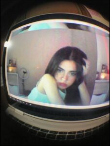 A fisheye lens image shows a young woman with long dark hair, wearing a light blue top, looking at a computer screen. She has her right hand touching her hair. The computer screen displays a blurred image of the woman and a room with furniture.