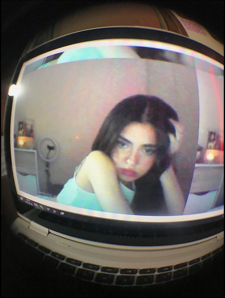 A fisheye lens image shows a young woman with long dark hair, wearing a light blue top, looking at a computer screen. She has her right hand touching her hair. The computer screen displays a blurred image of the woman and a room with furniture.