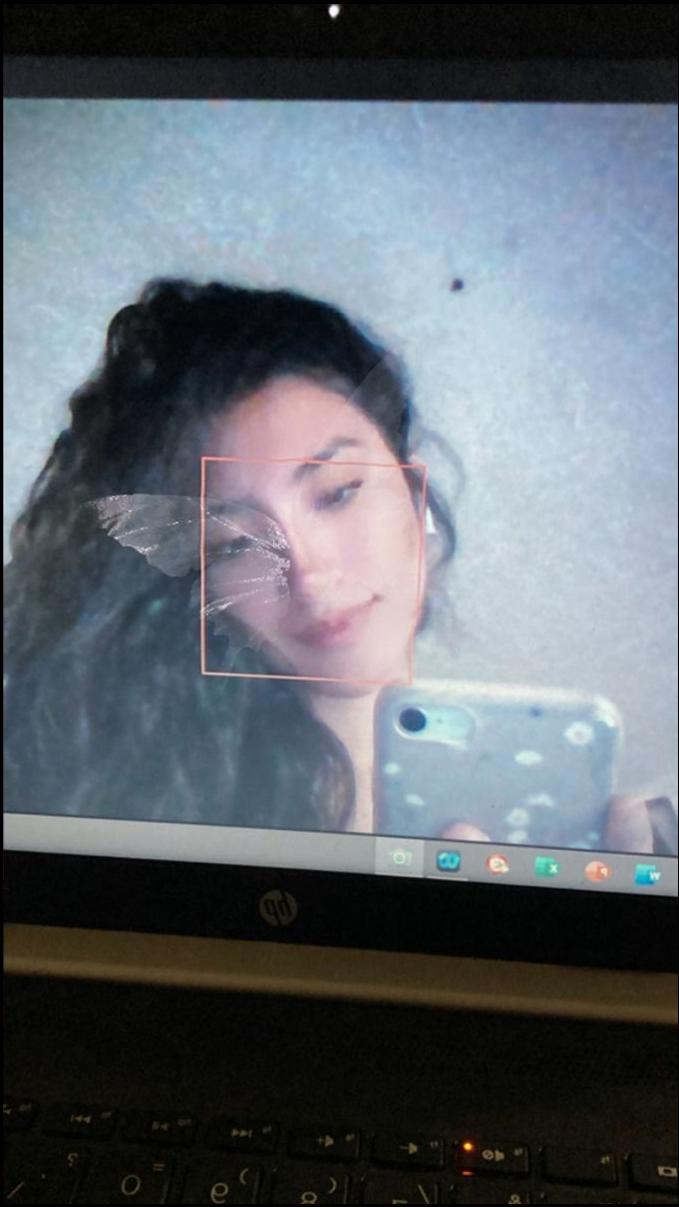 A selfie taken on a laptop screen shows a cracked or broken mirror reflecting a young woman with dark curly hair. The cracked area is highlighted by a red box on the mirror's reflection.