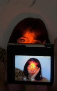 A person holds a digital camera displaying a close-up image of another person with reddish hair. The camera's LCD screen shows a red/orange glow around the subject's face. The camera's settings display "F8 0".