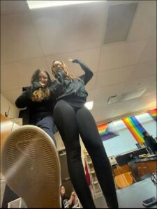 Two individuals stand in a classroom, one with dreadlocks and the other with raised arms. A person's foot is visible in the foreground, partially obscuring the view. Rainbow flags hang on the wall in the background.