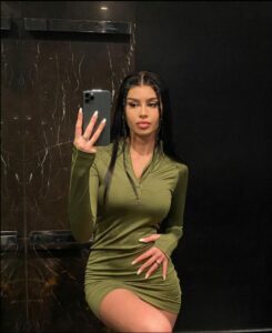 A woman with long dark hair, wearing a green zip-up dress, takes a selfie in a bathroom mirror. She holds a dark-colored smartphone in her right hand and wears a ring on her left ring finger. The bathroom features dark marble or stone walls.