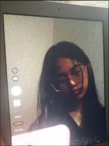A selfie taken on a tablet displays a person with long dark hair wearing glasses. They are positioned against a light-colored background with camera and video selection options visible in the upper left corner.
