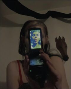 A person holds a flip phone displaying a selfie of a woman with a tongue sticking out. The phone is positioned in front of a wall with a dark object and a wooden horse decoration.