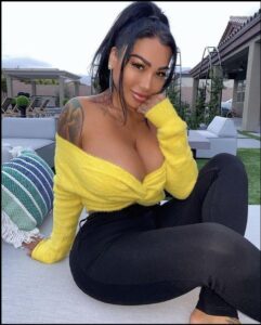 A woman with dark hair styled in a high ponytail sits outdoors, wearing a yellow off-the-shoulder top and black pants. She has tattoos on her left shoulder and arm and is wearing dangling earrings. A striped pillow and outdoor furniture are visible in the background.