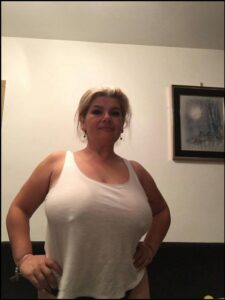 A woman with blonde hair, wearing a white tank top, stands with her hands on her hips against a white wall. A framed picture hangs on the wall behind her.