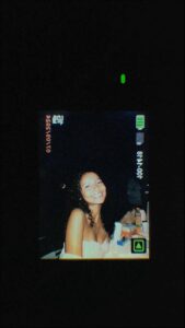 A video still displays a woman with curly hair, smiling and looking upwards. She is wearing a white garment and seated at a table with items on it. The video's timestamp reads "8:44-00:01". A green indicator light is visible in the upper right corner.