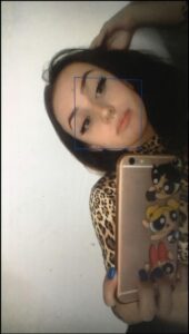 A person holds a phone displaying the Powerpuff Girls cartoon, wearing a leopard-print top. They are looking directly at the camera with a blue rectangular box superimposed on their face.