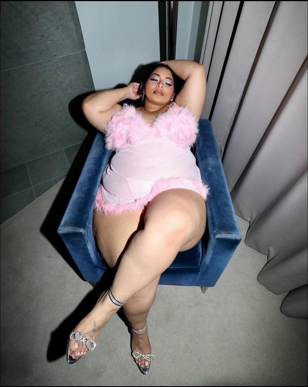 A woman lies on a blue velvet armchair, wearing a light pink, feathered bodysuit and silver high-heeled sandals. She rests her head on her left arm, with her right hand touching her hair. The background includes a tiled wall and a light-colored curtain.
