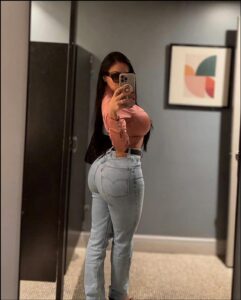 A woman takes a mirror selfie in a bathroom, wearing a pink cropped top and light blue jeans. She holds a smartphone in her right hand and wears sunglasses. A geometric abstract painting hangs on the wall behind her.