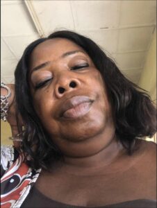 Close-up of a woman with dark hair, wearing a patterned top. Her eyes are partially closed, and she has a slight smile. A portion of a bracelet is visible on her left wrist. The background suggests an indoor setting with a light-colored ceiling.